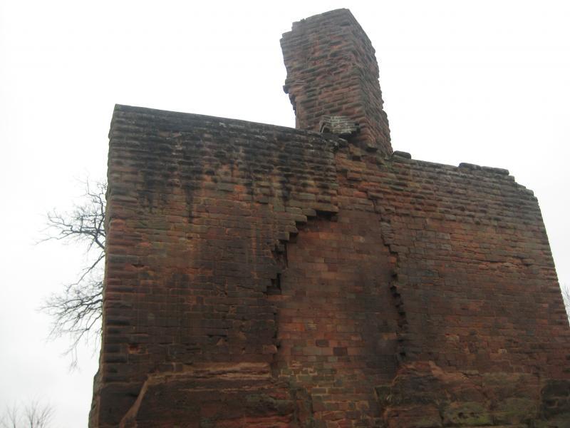 A weathered brick wall with a partially crumbling structure and a tall chimney protruding from the top, set against a cloudy sky. Some bare tree branches are visible nearby. Hohenecken Trails mountain bike trail.