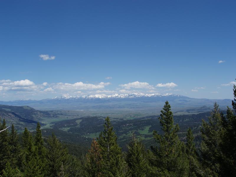A panoramic view of a mountain range under a clear blue sky, with scattered white clouds. In the foreground, there are green coniferous trees, while the expansive landscape features valleys and distant snow-capped peaks. Grassy Mountain Loop mountain bike trail.