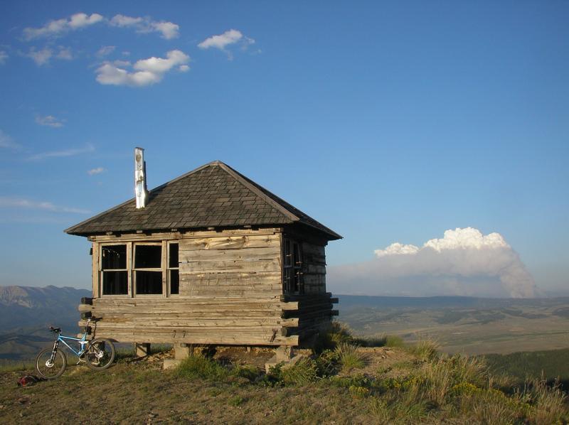 A rustic wooden cabin sits on a hillside, with a bicycle parked nearby. In the background, mountains stretch into the distance beneath a blue sky, with white clouds and a plume of smoke visible on the horizon, suggesting a fire. The scene conveys a sense of solitude and natural beauty. Monument Ridge mountain bike trail.