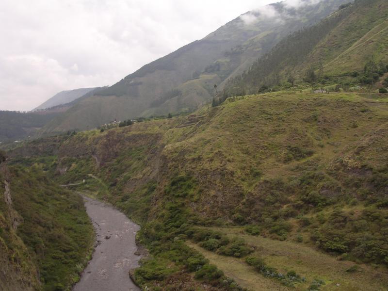 A scenic landscape featuring a winding river flowing through a valley, surrounded by lush green hills and mountains. The sky is partly cloudy, adding a sense of tranquility to the natural setting. El Triunfo Loop mountain bike trail.