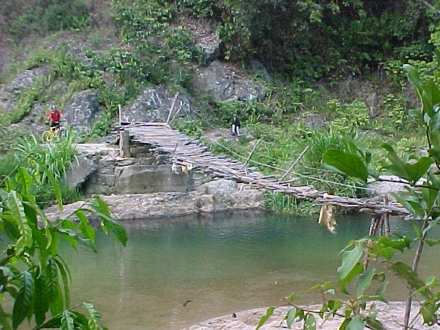 A rustic wooden bridge made of bamboo spans a shallow, clear pond surrounded by lush green vegetation and rocky terrain. Two individuals are seen near the bridge—one is approaching from the left, while the other stands on the opposite side. The serene natural setting features dense greenery and hillside, creating a peaceful outdoor atmosphere. Arroyaso - La Sal mountain bike trail.