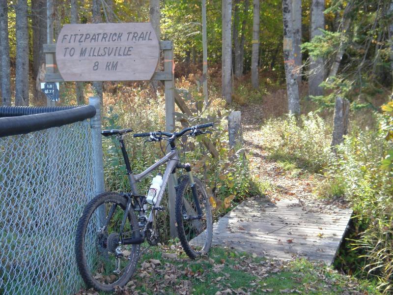 A mountain bike is parked next to a sign that reads "Fitzpatrick Trail to Millsville 8 km." The scene is set in a wooded area with autumn foliage, featuring a wooden pathway leading into the trail. A chain-link fence is visible in the foreground. Fitzpatrick Trail mountain bike trail.