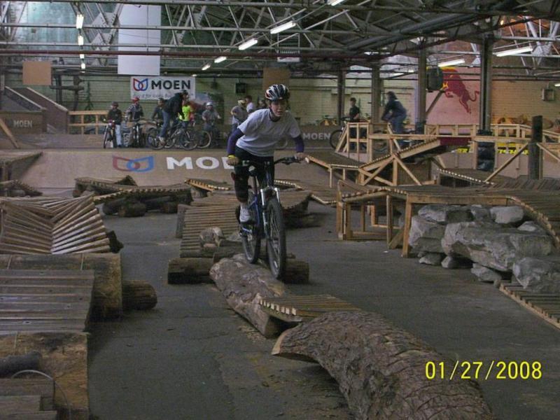 A person riding a mountain bike skillfully navigates a wooden obstacle course in an indoor biking facility. The scene includes various ramps and structures, with additional riders in the background. The atmosphere reflects a competitive and active biking environment. Rays Indoor Mtb Park mountain bike trail.