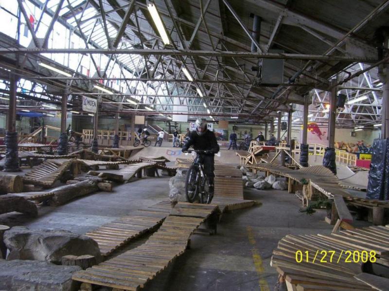 A rider in protective gear is navigating a wooden obstacle course inside a large indoor BMX park, featuring various ramps, logs, and rocks. Other cyclists are visible in the background, and the facility has high ceilings and industrial-style architecture. Rays Indoor Mtb Park mountain bike trail.