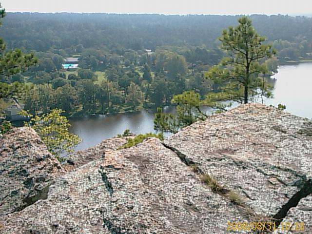 A rocky outcrop overlooking a serene lake surrounded by lush greenery, including trees and residential areas, under a clear sky. Robbers Cave State Park mountain bike trail.
