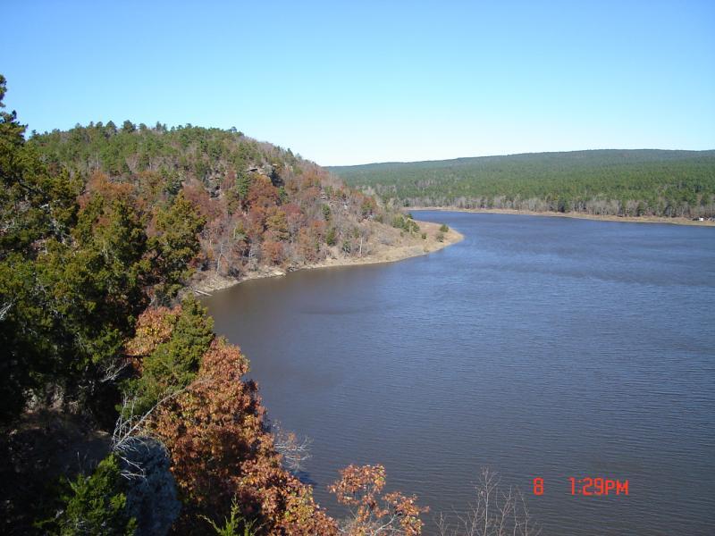 A panoramic view of a winding river surrounded by lush green hills and autumn foliage under a clear blue sky. Robbers Cave State Park mountain bike trail.