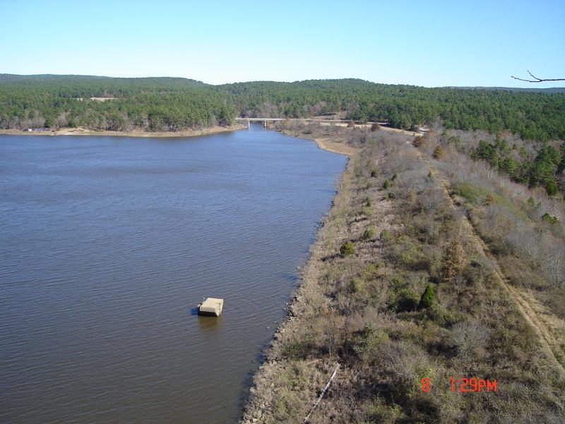 A panoramic view of a calm lake bordered by trees and a gentle shoreline. In the foreground, a small dock extends into the water. In the background, rolling hills covered in greenery stretch under a clear blue sky. The scene conveys a tranquil natural setting. Robbers Cave State Park mountain bike trail.
