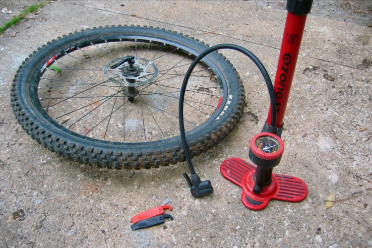 A mountain bike tire lying on the ground next to a red hand pump. The pump features a gauge on top, and two tire levers in red and black are placed nearby. The surface beneath is concrete with some dirt and small debris.
