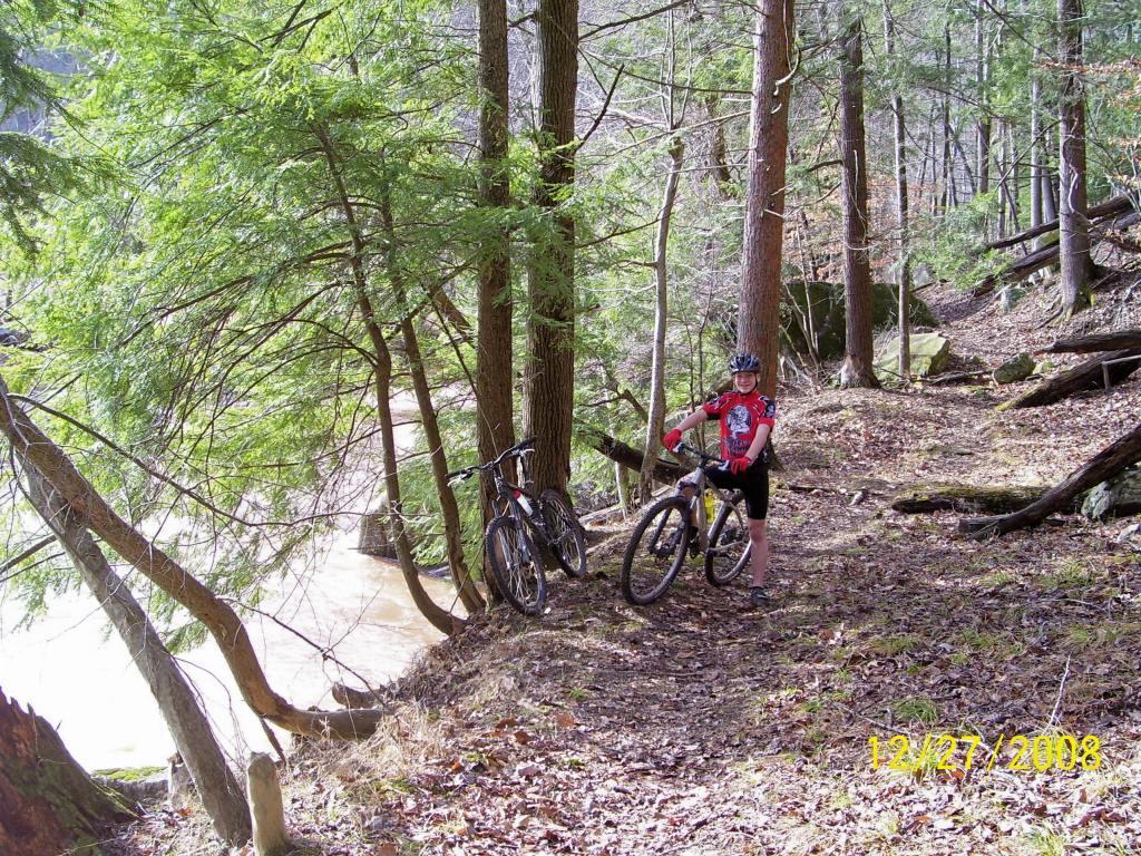A person wearing a red cycling jersey and shorts stands next to two mountain bikes on a forested path. Lush green trees surround the area, and a river can be seen in the background. The scene captures a moment of outdoor adventure. North Bend State Park mountain bike trail.