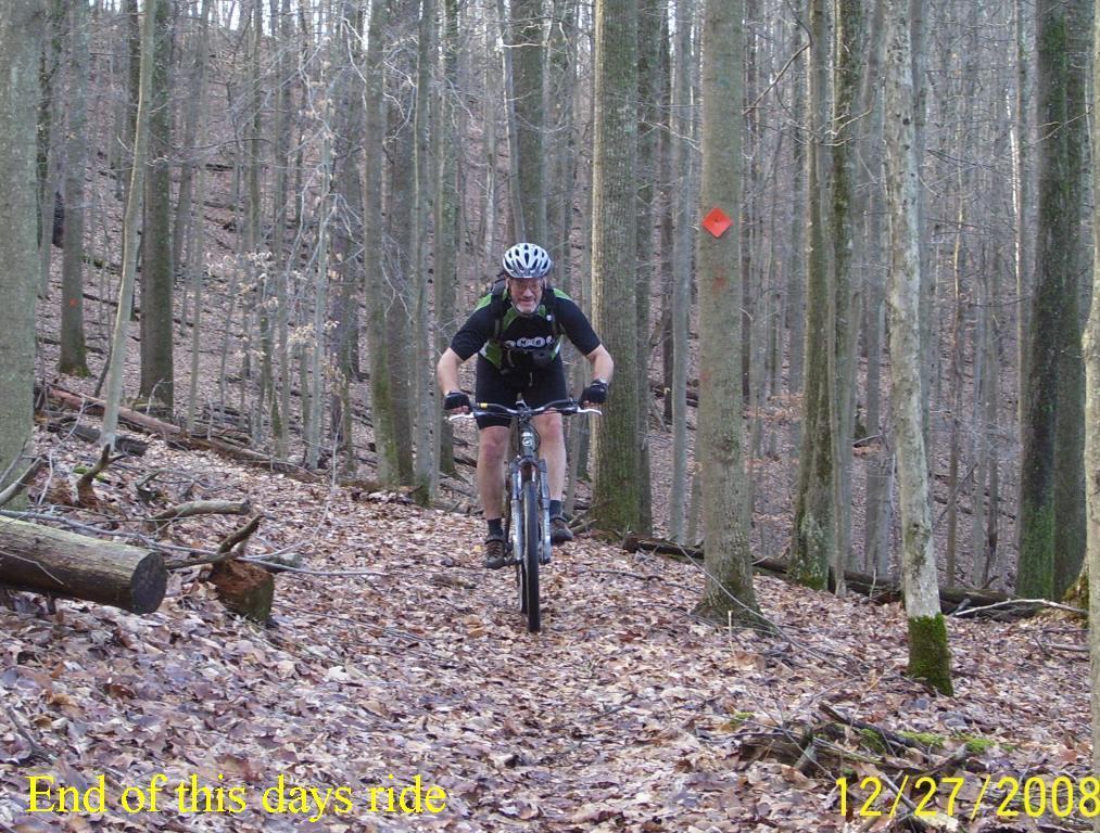 A mountain biker navigates a trail through a forest of bare trees and fallen leaves, with a small orange trail marker visible in the background. The scene captures the natural beauty of winter hiking and biking, emphasizing the winding path and the rider's focus. The date "12/27/2008" is displayed in the corner, marking the occasion. North Bend State Park mountain bike trail.