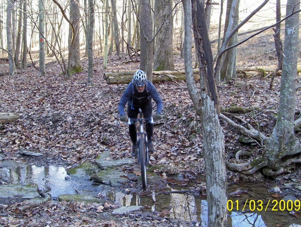 A mountain biker navigating through a wooded area, riding over rocks and a small stream, with fallen leaves covering the ground and trees in the background. The photo is taken in a natural setting, showcasing outdoor cycling activity. North Bend State Park mountain bike trail.
