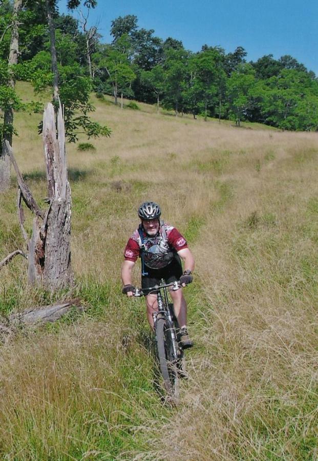 A person riding a mountain bike through a grassy field, with trees and a clear blue sky in the background. The cyclist is wearing a helmet and a dark red jersey, smiling as they navigate a dirt path surrounded by tall grass. North Bend State Park mountain bike trail.