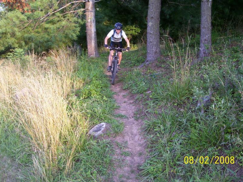 A mountain biker navigates a narrow dirt trail surrounded by tall grass and trees, wearing a helmet and a backpack. The scene captures the outdoor adventure spirit in a lush, green environment. City Trails - Marietta Hs mountain bike trail.