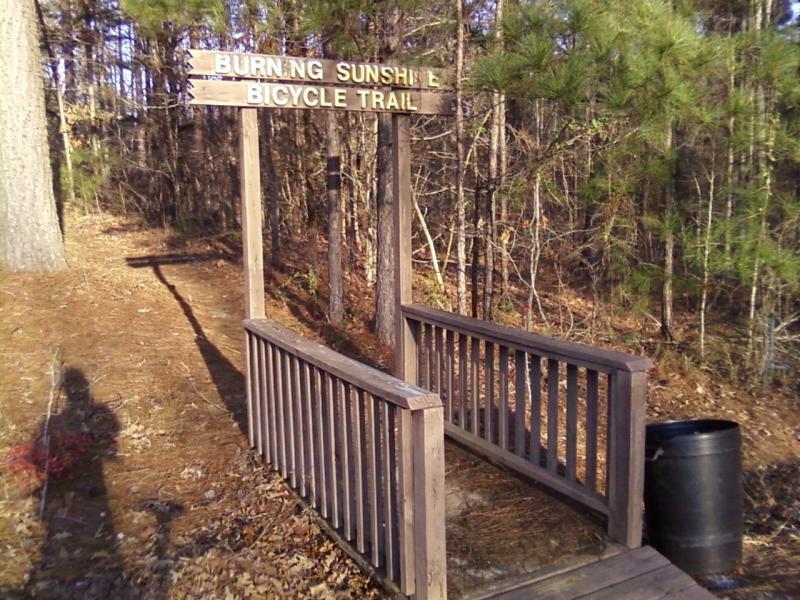 Wooden sign marking the entrance to the Burning Sunlight Bicycle Trail, surrounded by trees and foliage, with a small bridge leading into the trail. A black barrel is visible nearby. Burning Sunshine mountain bike trail.