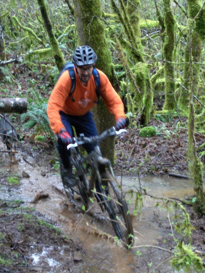 A person riding a mountain bike through a muddy trail in a dense forest. The rider, wearing an orange long-sleeve shirt, a helmet, and gloves, is navigating through stagnant water and mud, surrounded by lush green trees and moss. McDonald / Dunn Forests mountain bike trail.