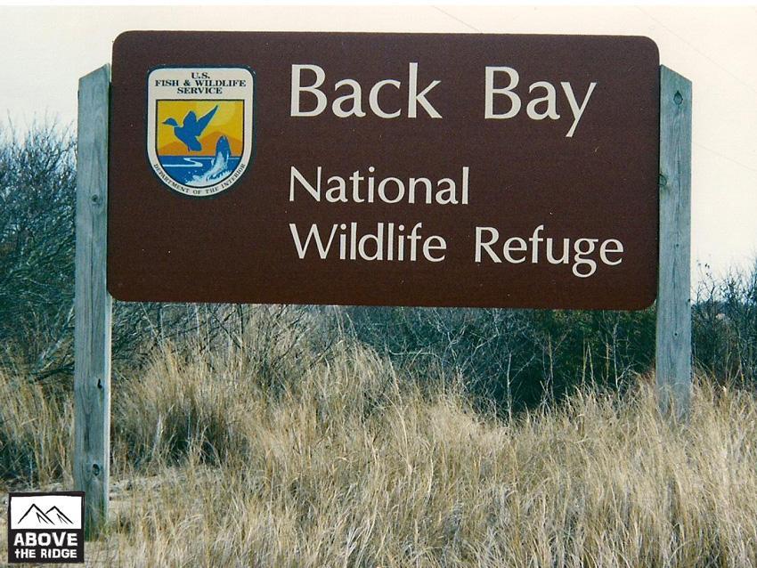Sign for Back Bay National Wildlife Refuge, featuring the U.S. Fish & Wildlife Service logo. The sign is located in a grassy area, indicating the entrance to the refuge. False Cape State Park (back Bay Wildlife Refuge) mountain bike trail.