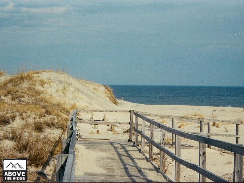 A wooden boardwalk leads through sandy dunes towards a calm ocean under a partly cloudy sky. Grasses grow alongside the path, emphasizing the natural coastal landscape. False Cape State Park (back Bay Wildlife Refuge) mountain bike trail.