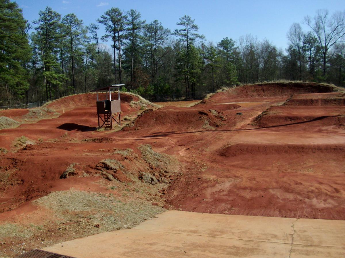 An empty dirt bike track featuring various jumps and mounds, with a small observation tower on the left. The area is surrounded by trees under a clear blue sky. The ground is composed of reddish dirt and some grassy patches. East Macon Park mountain bike trail.