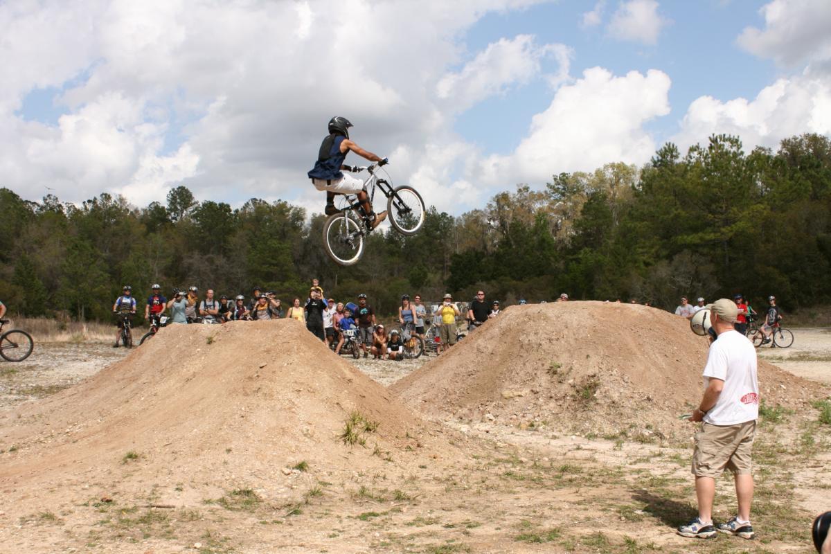 A BMX rider performing a jump over a dirt ramp during a biking event, with an audience of spectators in the background, some on bicycles. The scene is set outdoors with a mix of clouds and trees in the background. Santos mountain bike trail.