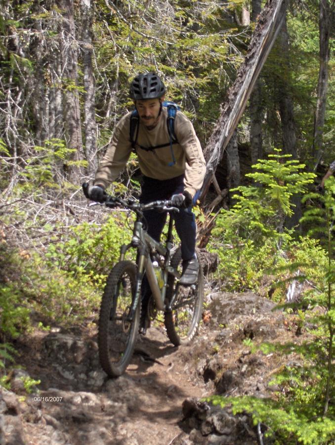 A person riding a mountain bike on a rocky trail surrounded by trees and greenery, wearing a helmet and backpack. Mckenzie River Trail mountain bike trail.