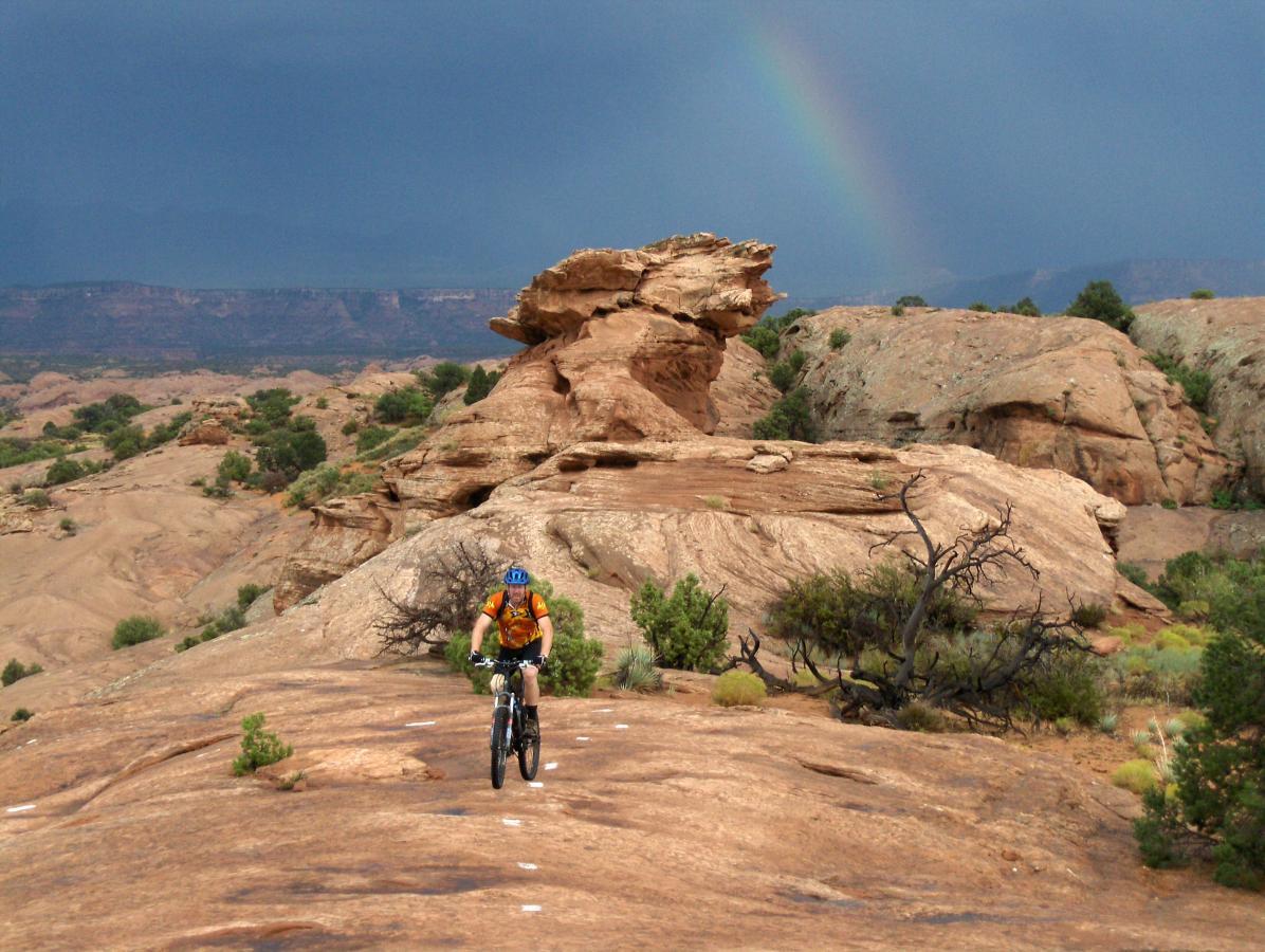 A mountain biker rides along a rocky trail surrounded by desert terrain, with a rainbow visible against a dark, cloudy sky in the background. The landscape features unique rock formations and sparse vegetation. Slickrock mountain bike trail.