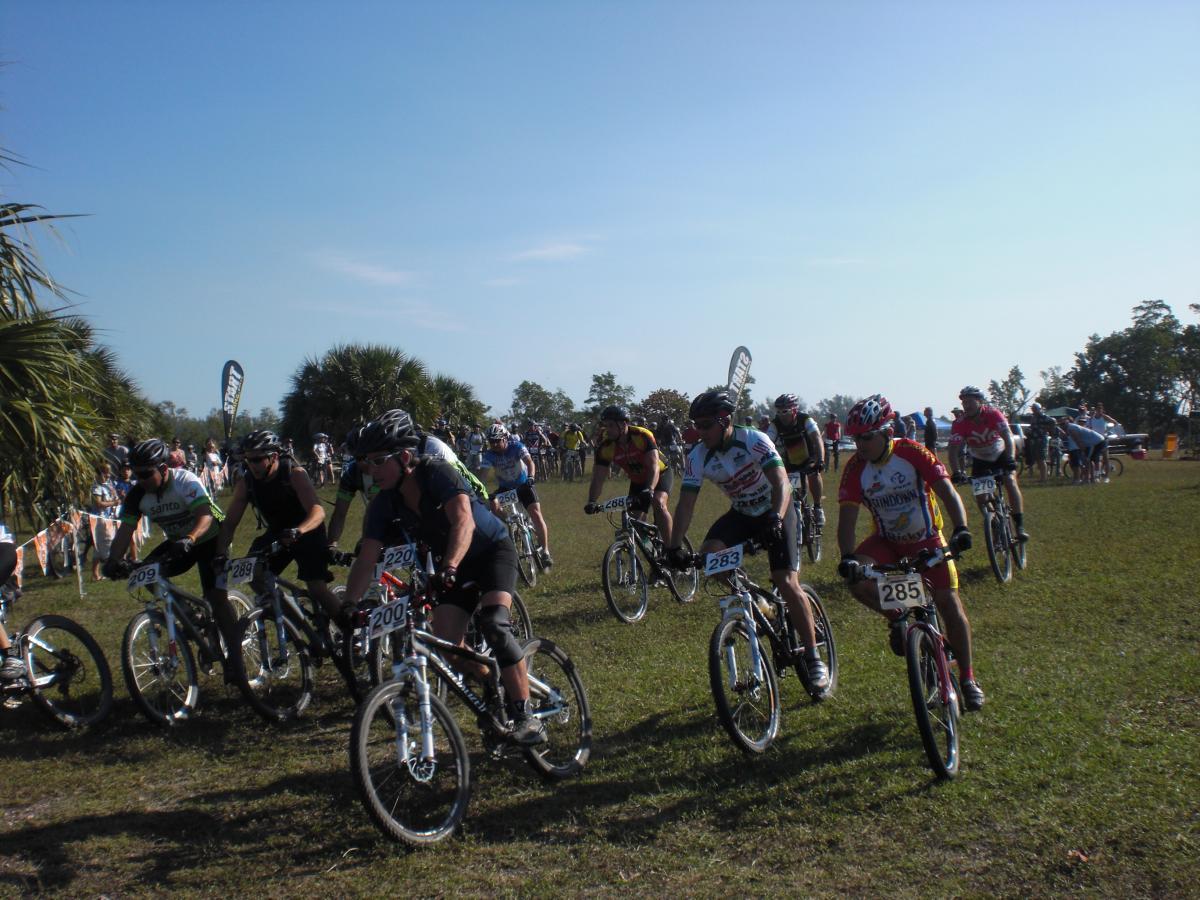 A group of mountain bikers lined up at the start of a race, wearing numbered jerseys and helmets. The scene takes place on a grassy field with spectators in the background and palm trees dotting the landscape under a clear blue sky. Oleta River State Park mountain bike trail.