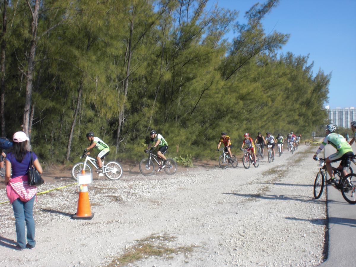 A group of mountain bikers competes in a race on a gravel path surrounded by tall trees, with a spectator watching on the left. Traffic cones indicate the race route. The sky is clear and blue, creating a vibrant outdoor atmosphere. Oleta River State Park mountain bike trail.