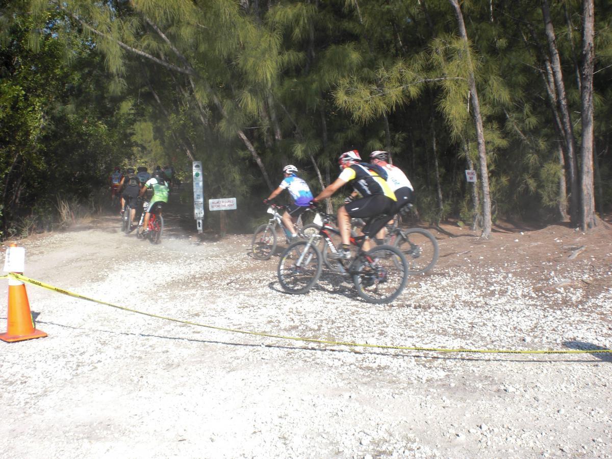 A group of mountain bikers riding along a gravel path bordered by trees. The cyclists, dressed in colorful jerseys and helmets, are kicking up dust as they navigate a terrain marked by a signpost and caution tape. The scene captures the energy and movement of the riders in a natural outdoor setting. Oleta River State Park mountain bike trail.