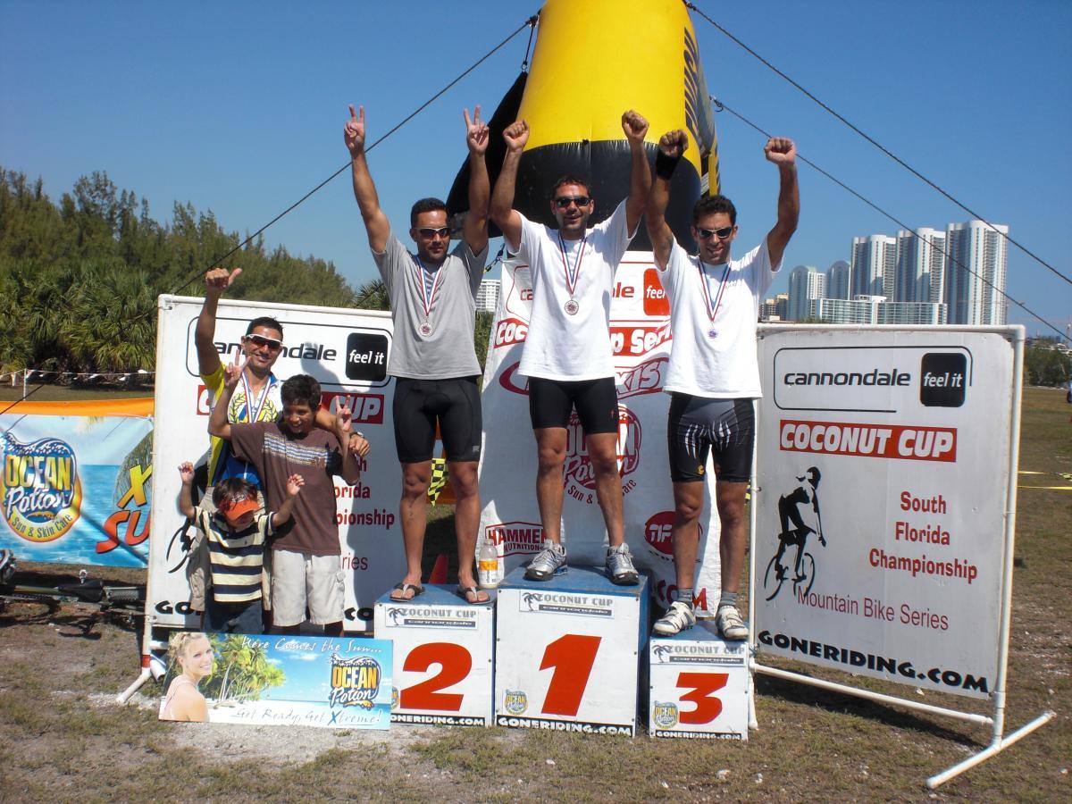A group of four men stands on a podium celebrating their medals at the Coconut Cup South Florida Championship mountain bike race. The first-place winner is on the far left with a gold medal, followed by the second-place winner, who holds up two fingers, and the third-place winner, who raises his arms in excitement. Two young boys stand in front of them, all joyfully showing their enthusiasm. The backdrop features a clear sky and palm trees, suggesting a sunny outdoor setting. Oleta River State Park mountain bike trail.