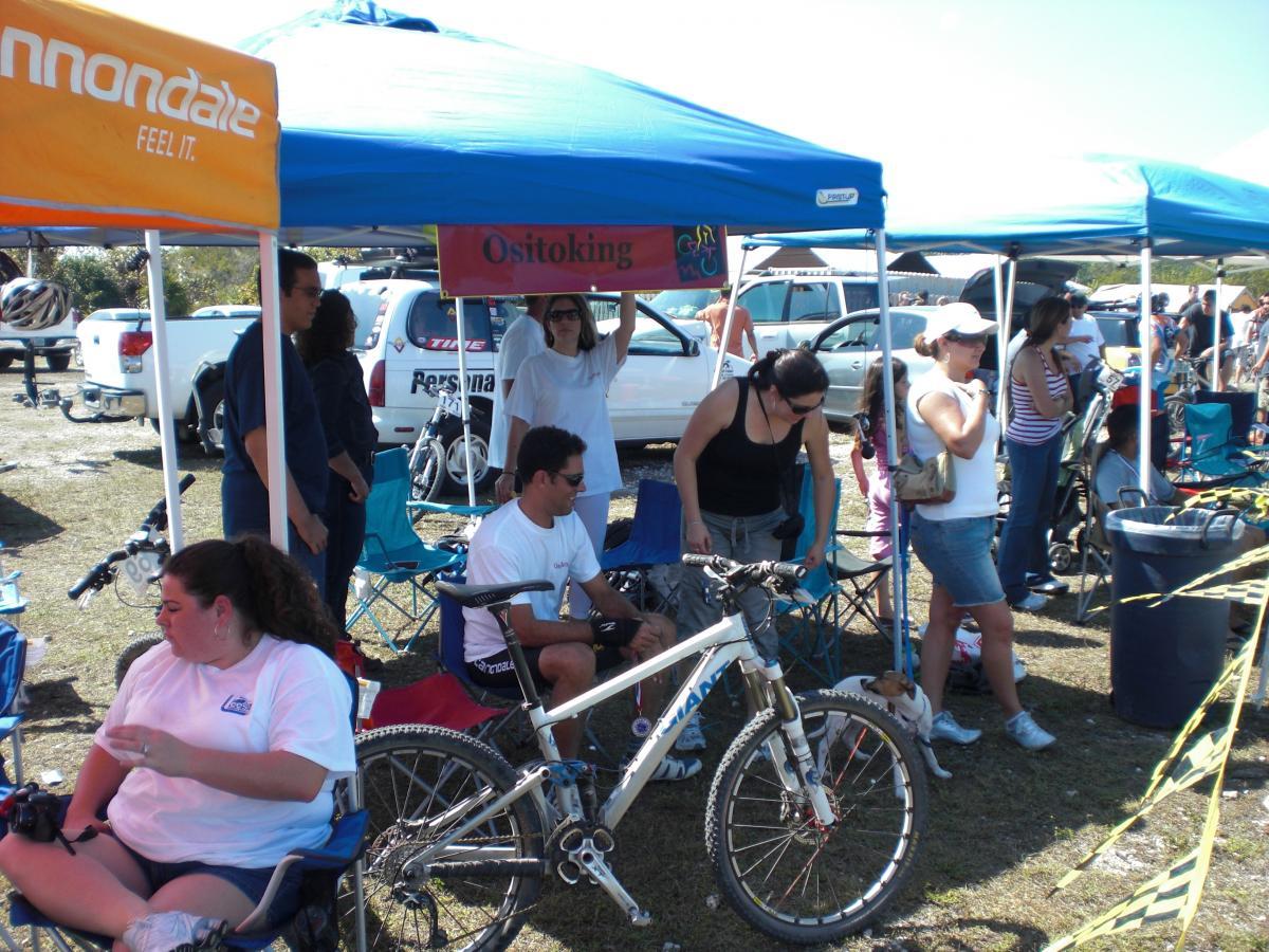 A group of people gathered under blue tents at an outdoor event, with bikes parked nearby. Some individuals are sitting in folding chairs, while others are standing and chatting. A banner labeled "Ositoking" is visible above one tent, and there are bicycles in the foreground and background. The scene is lively, with sunny weather and various vehicles parked in the distance. Oleta River State Park mountain bike trail.