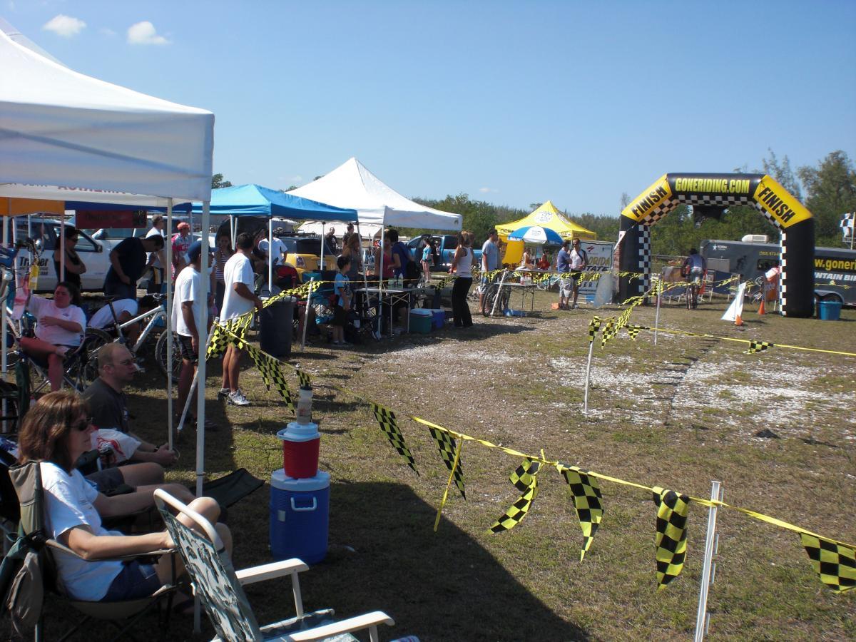 A busy outdoor event scene featuring tents and spectators gathered near a finish line. In the foreground, people relax in chairs while others stand and interact. The finish line is marked by a prominent inflatable arch with "goneriding.com FINISH" signage, surrounded by caution tape. Various tents in the background showcase vendors and participants, set against a clear blue sky. Oleta River State Park mountain bike trail.
