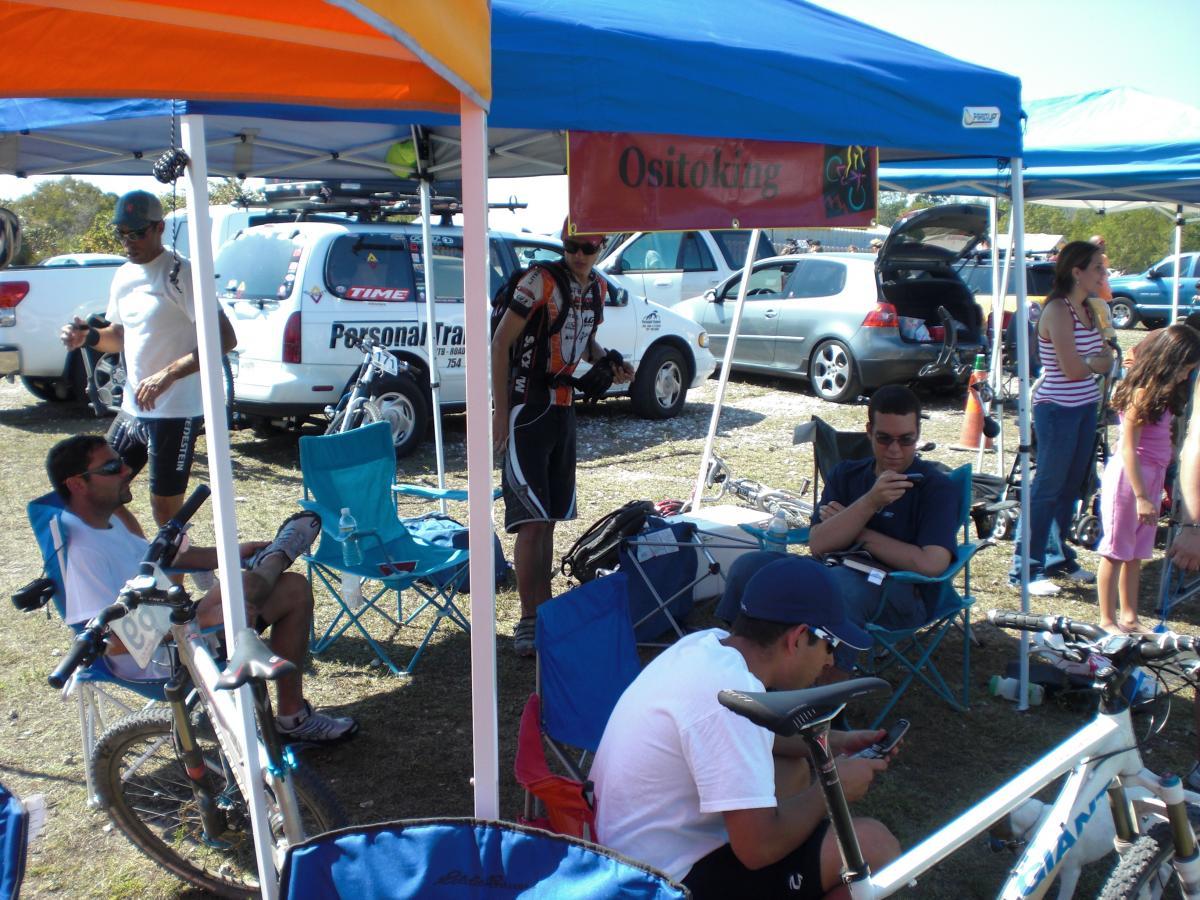A group of people enjoying a sunny day at a biking event, sitting under colorful tents in a parking area. Some individuals are relaxing in folding chairs, while others are engaging with their smartphones or chatting. Bicycles are parked nearby, and there are vehicles in the background. The setting appears casual and social, reflective of an outdoor gathering focused on biking. Oleta River State Park mountain bike trail.