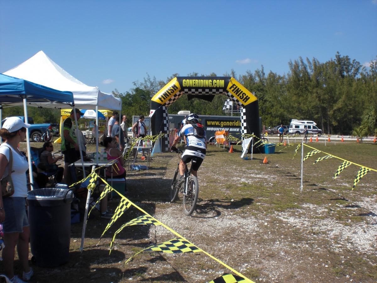 A mountain biker crosses the finish line under an inflatable archway marked "FINISH" at a bike race event. Spectators and participants are gathered around, with tents and banners visible in the background. The ground is slightly uneven, with yellow and black checkered tape marking the path. Oleta River State Park mountain bike trail.