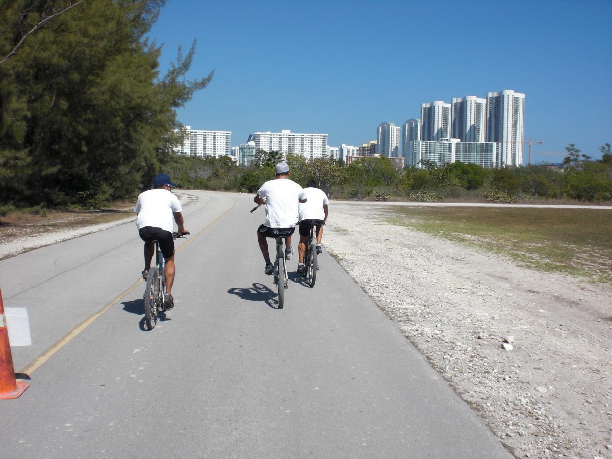 Three cyclists in white t-shirts ride along a paved path bordered by greenery, with a city skyline in the background under a clear blue sky. Oleta River State Park mountain bike trail.