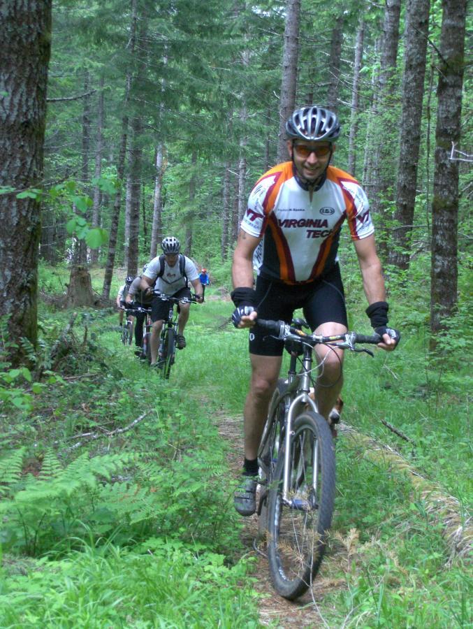 Two mountain bikers riding through a dense forest trail, surrounded by tall trees and greenery. The front cyclist wears a brightly colored jersey and a helmet, smiling while navigating the path, while a second cyclist follows closely behind, dressed in a white jersey. The scene captures the essence of outdoor adventure and exercise. McDonald / Dunn Forests mountain bike trail.
