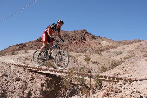 A mountain biker riding over a narrow wooden bridge in a rocky desert landscape, with rugged hills and a clear blue sky in the background. Bootleg Canyon mountain bike trail.