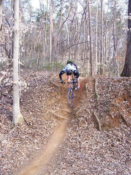 A mountain biker in mid-air jumps off a dirt ramp on a narrow trail surrounded by trees and fallen leaves. USNWC mountain bike trail.
