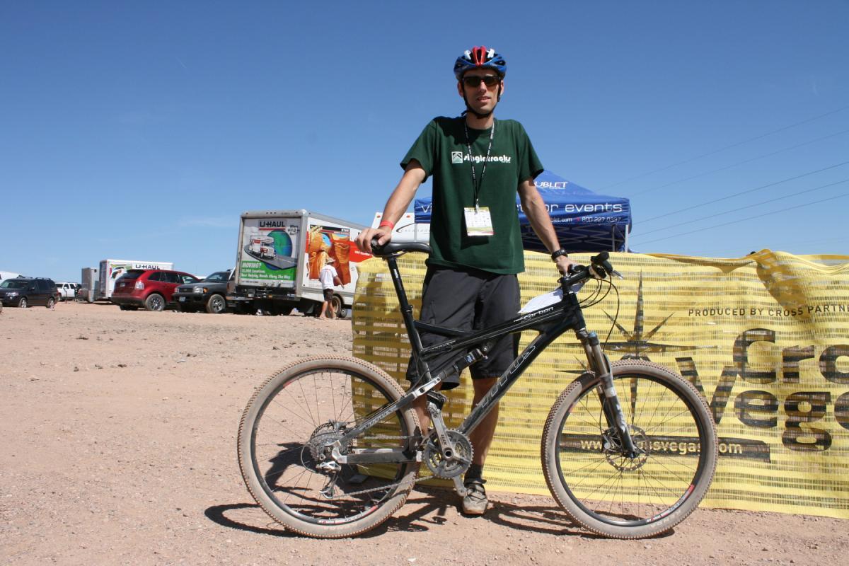 A person wearing a green t-shirt and a helmet stands next to a mountain bike in a desert-like area. In the background, there are parked vehicles and a yellow banner with text. The sky is clear and blue, indicating a sunny day. Bootleg Canyon mountain bike trail.