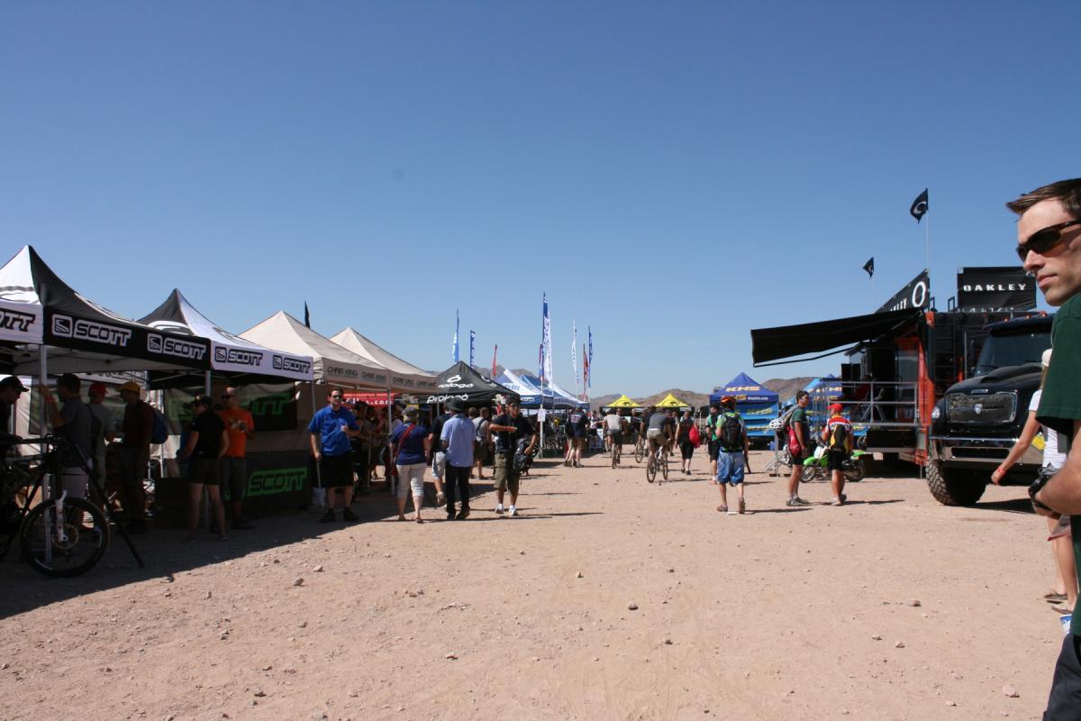 A busy outdoor event scene featuring various vendor tents, including brands like Scott and Oakley, where attendees are browsing and interacting. People of diverse ages are seen walking along a dusty path, with bicycles and promotional flags visible in the background under a clear blue sky. Bootleg Canyon mountain bike trail.