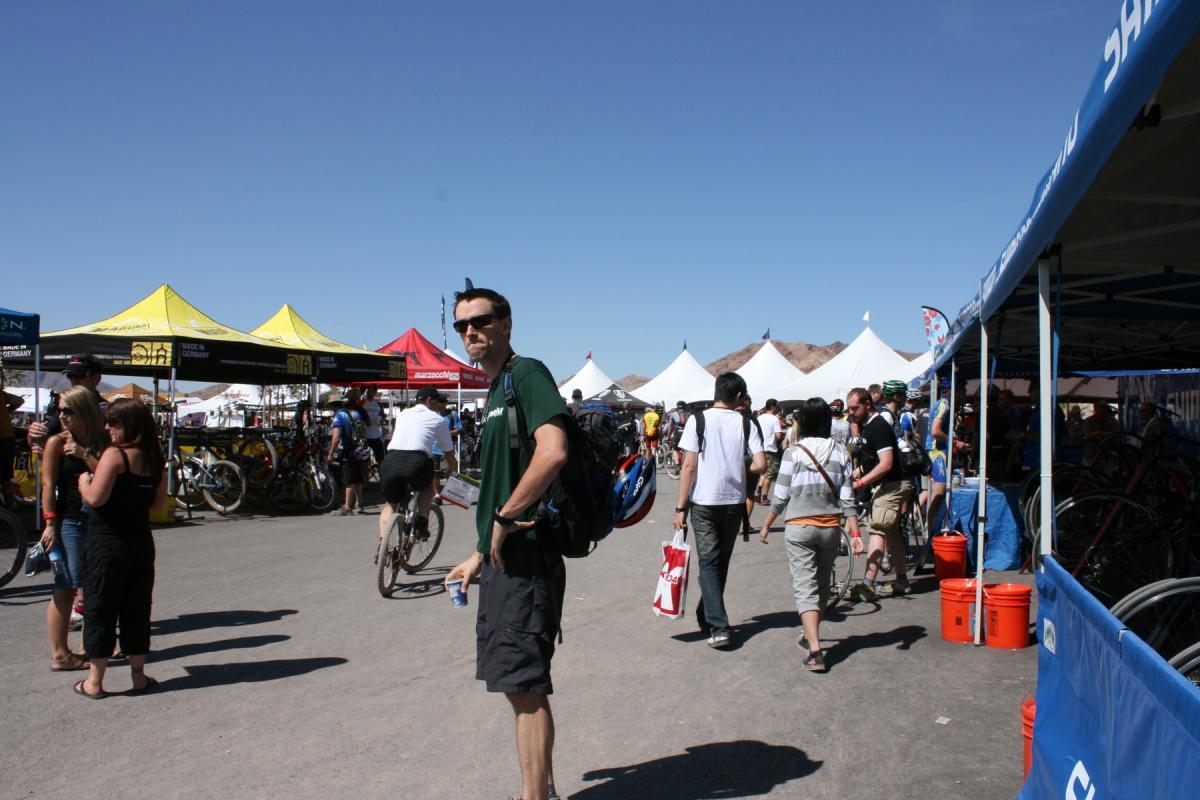 A busy outdoor event with numerous people milling about, surrounded by colorful tents displaying bicycles and cycling gear. In the foreground, a man wearing sunglasses and a backpack stands with a drink in hand, looking towards the camera. Various attendees are engaged in conversation and browsing the booths, with a clear blue sky above them. Bootleg Canyon mountain bike trail.