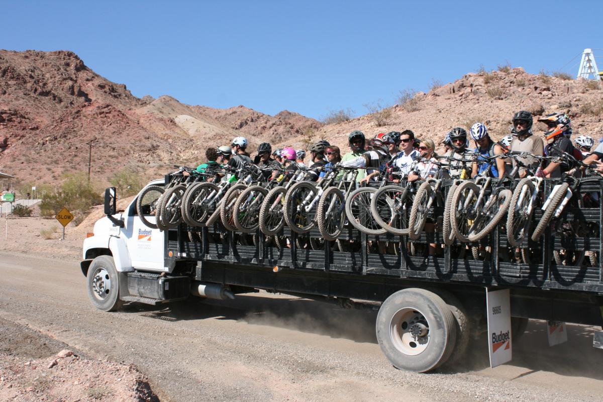 A group of mountain bikers sitting on a flatbed truck, with their bicycles secured in racks behind them. The truck is on a dirt road surrounded by rocky landscapes and mountains under a clear blue sky. Dust is being kicked up from the road as the truck moves. Bootleg Canyon mountain bike trail.