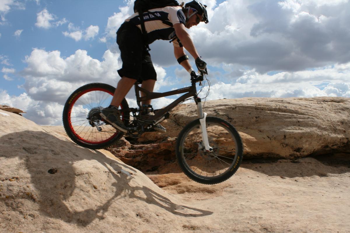 A mountain biker jumps over a rocky terrain, captured mid-air with a clear blue sky and fluffy clouds in the background. The rider wears a helmet and protective gear, showcasing an athletic pose as the bike's wheels are off the ground. Gooseberry Mesa mountain bike trail.
