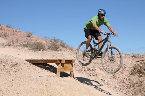 A cyclist in a green shirt and helmet performs a jump off a wooden ramp on a dirt trail, surrounded by a rocky landscape and sparse vegetation under a clear blue sky. Bootleg Canyon mountain bike trail.