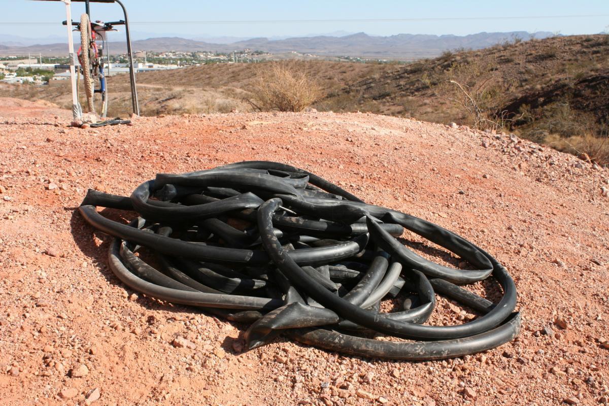 A pile of coiled black rubber hoses resting on a dry, reddish-brown terrain, with a distant view of a small town and hills in the background under a clear sky. Bootleg Canyon mountain bike trail.