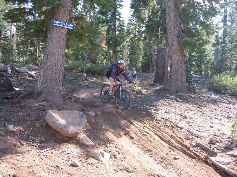 A mountain biker rides down a dirt path in a forest, with trees surrounding the area. A sign on a nearby tree indicates "SERVICE ROAD (NO BIKING ALLOWED)." Another cyclist is visible in the background, navigating the terrain. Northstar Bike Park mountain bike trail.