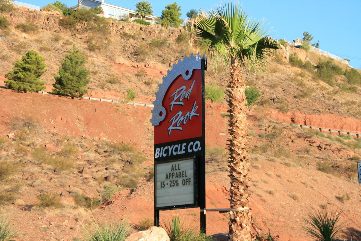 A roadside sign for "Red Rock Bicycle Co." displaying a promotion for all apparel with discounts ranging from 15% to 25% off. The sign is set against a backdrop of red rocky terrain and greenery, including palm trees.