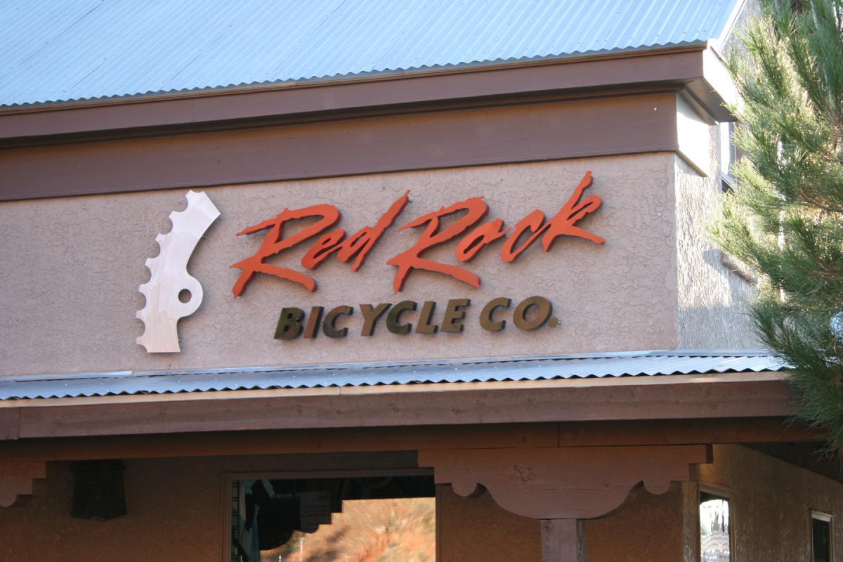 Signage for "Red Rock Bicycle Co." displayed on a building exterior, featuring a stylized bike gear graphic and bold lettering. The colors include red for "Red Rock" and black for "Bicycle Co." The setting includes part of a roof and greenery on the side.