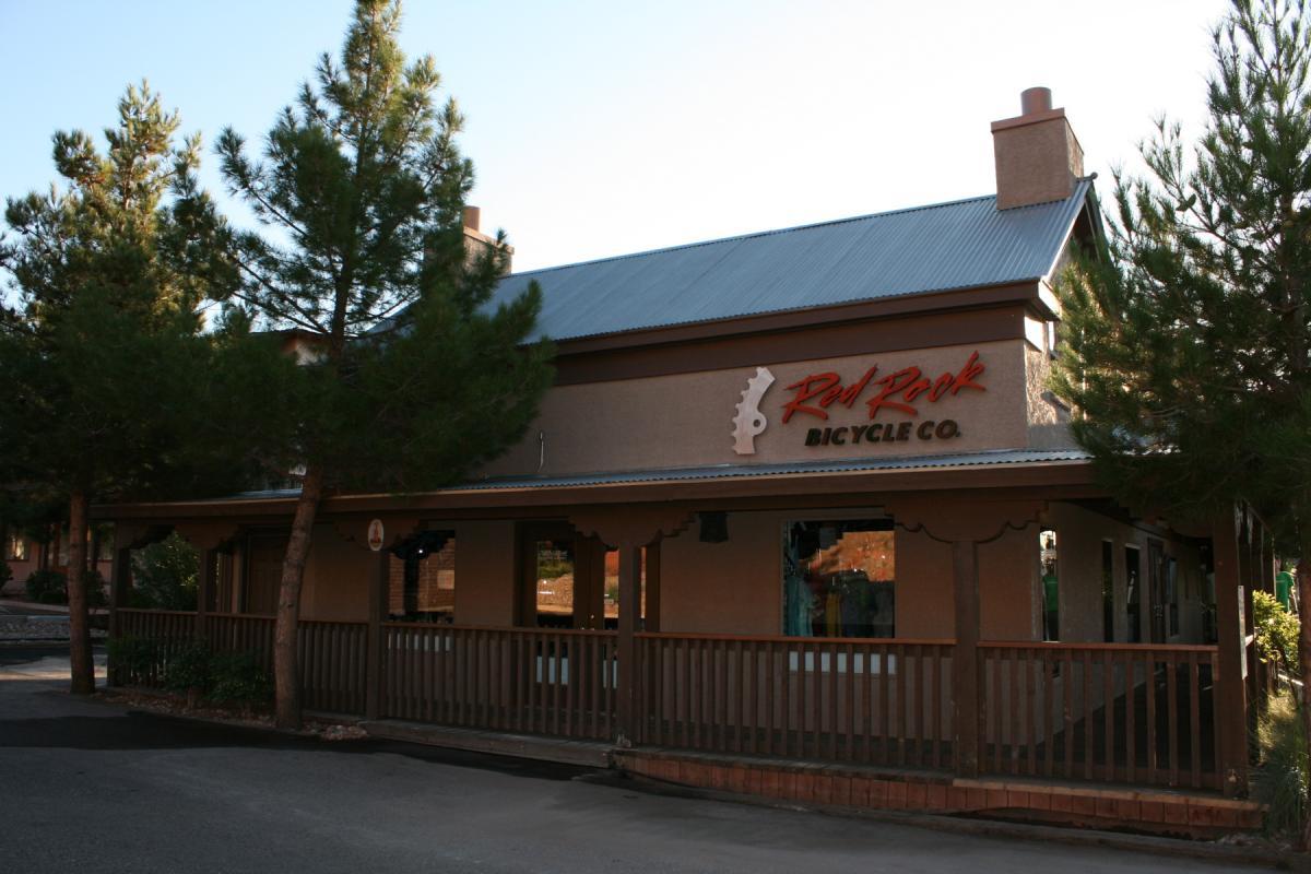 A bicycle shop with a wooden porch and a sign that reads "Red Rock Bicycle Co." surrounded by pine trees. The building features a sloped metal roof and large windows showcasing various items inside. The scene is set in a sunny outdoor environment.