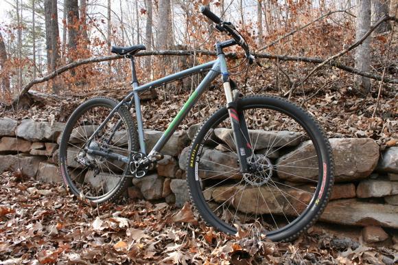 A mountain bike resting against a stone wall in a wooded area, surrounded by fallen leaves. The bike features a blue and green frame with thick tires, suitable for off-road terrain. The trees in the background have sparse foliage, indicating a season without many leaves. Yellow River mountain bike trail.