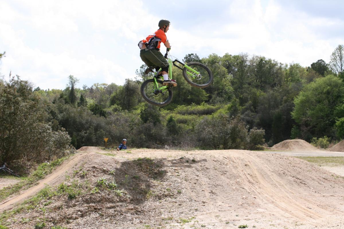 A young cyclist performs a jump on a mountain bike at a dirt track, soaring above a dirt ramp with lush greenery in the background. Another cyclist can be seen in the lower left, watching the jump. The sky is partly cloudy, suggesting a bright day for biking. Santos mountain bike trail.